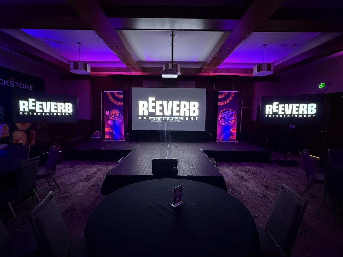 Three-screen corporate stage with the Reeverb Entertainment logo in a dark ballroom